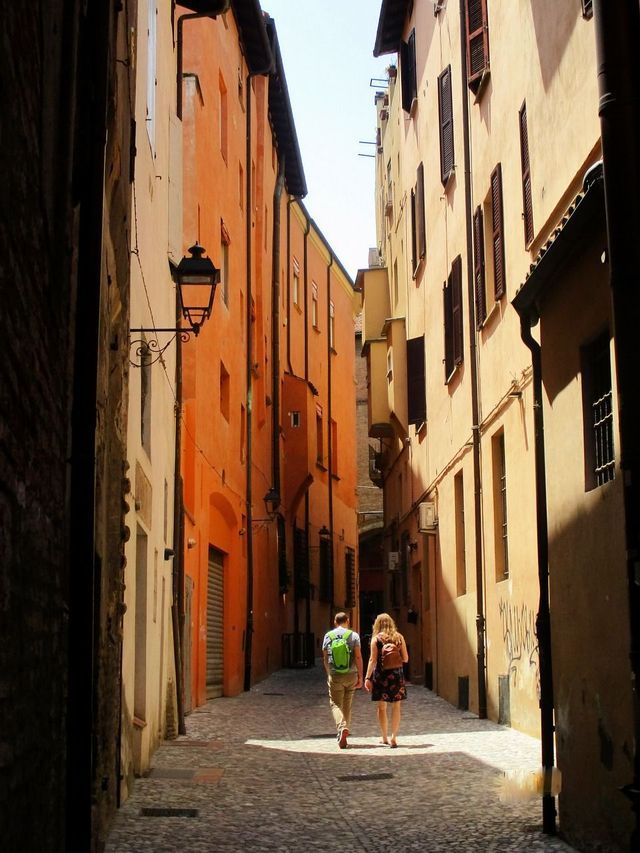 Un uomo e una donna con zaini camminano lungo uno stretto vicolo di ciottoli illuminato dal sole, tra alti edifici colorati.