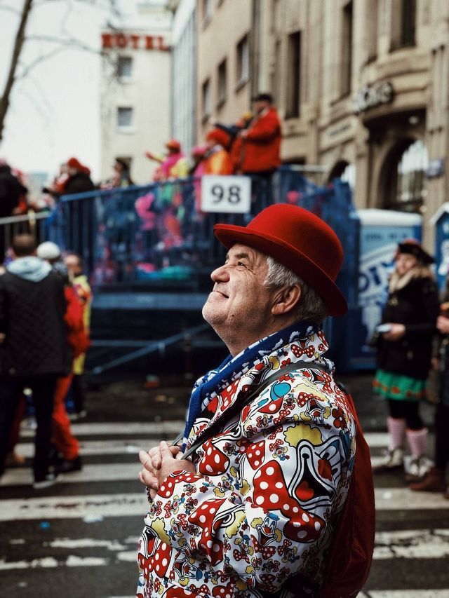 Un uomo anziano con una bombetta rossa e una giacca colorata con stampa a funghi che sorride durante una parata di strada.