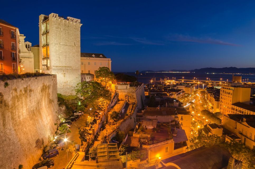 An ancient stone tower and walls overlooking a coastal city with illuminated streets at night.