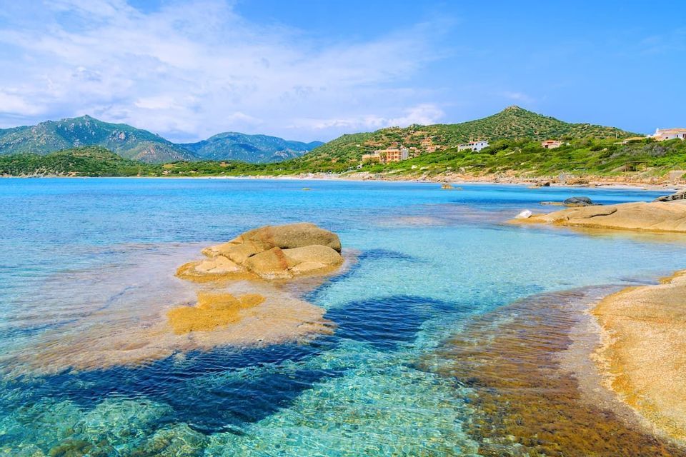 Large, smooth rocks sit in clear, shallow turquoise water with a sandy beach and green hills in the distance under a blue sky.