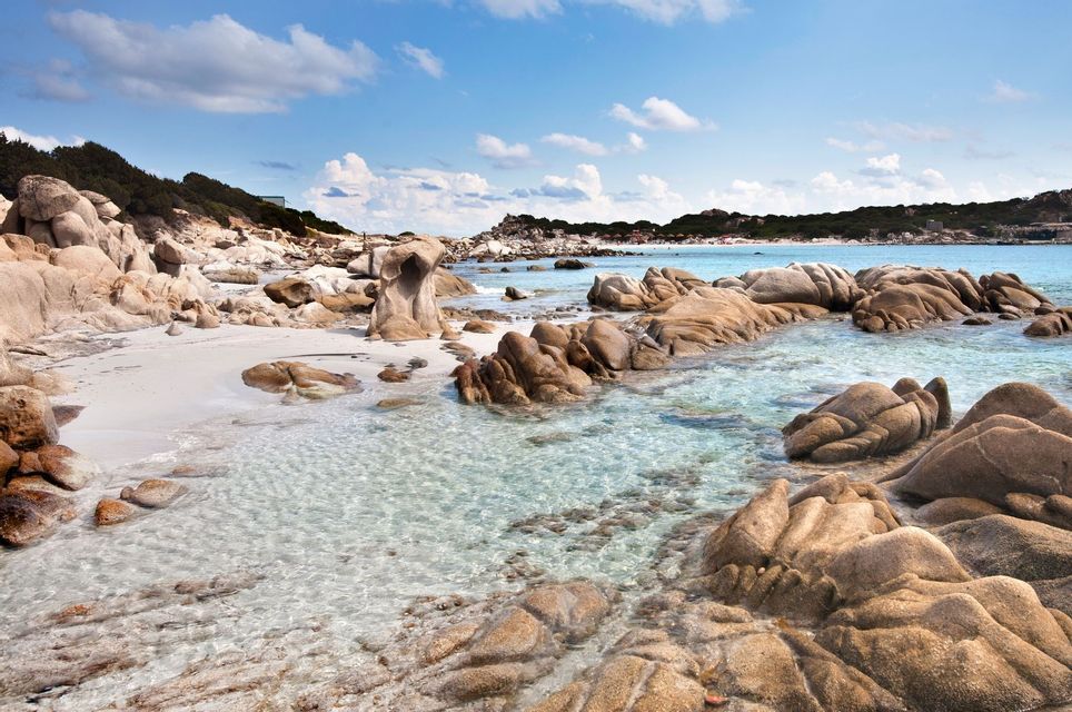 Large, smooth rocks are scattered on a white sand beach and in clear, shallow water under a blue sky with white clouds.