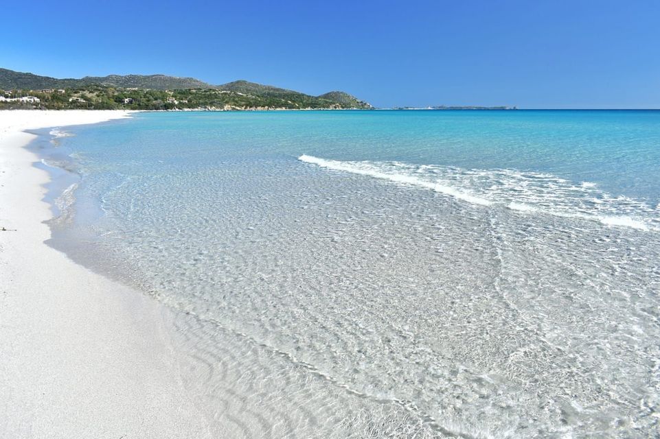 A white sand beach meets clear, turquoise water with gentle waves, set against a backdrop of green hills under a bright blue sky.