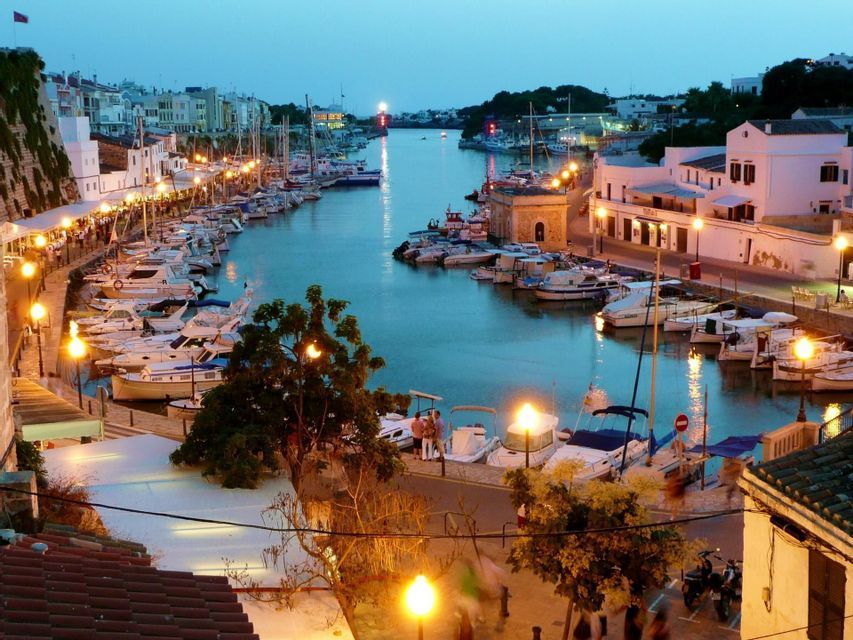 An evening view of a harbor filled with moored boats, where waterfront buildings and streetlights cast a warm glow on the water.