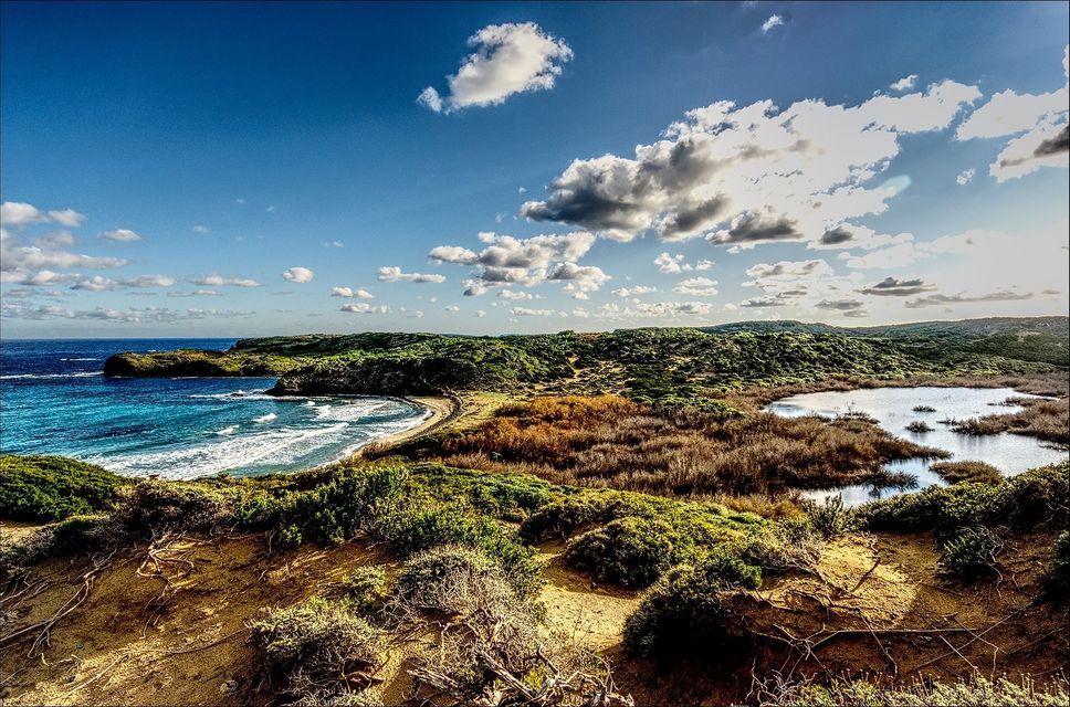 A panoramic view of a coastal landscape with a sandy bay, a small lagoon, and lush green hills under a partly cloudy blue sky.