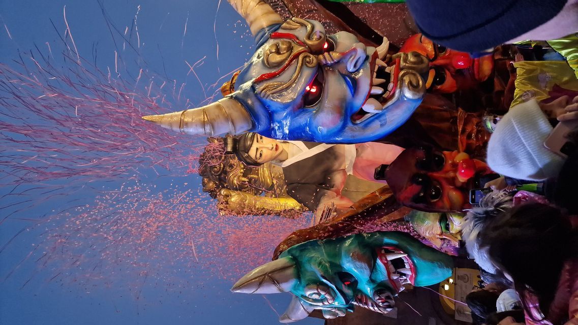 Inquadratura verticale dal basso di un grande carro di carnevale con maschere di demoni colorate e coriandoli rosa che esplodono nel cielo.
