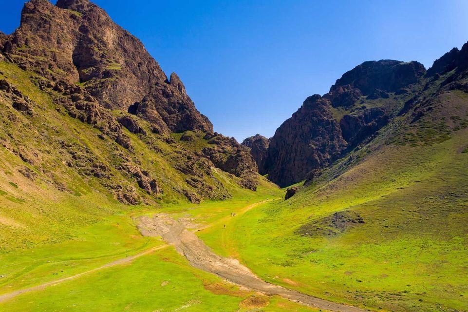 Un viaggio di gruppo WeRoad percorre un sentiero in un'ampia valle verde tra montagne rocciose, sotto un cielo azzurro limpido.