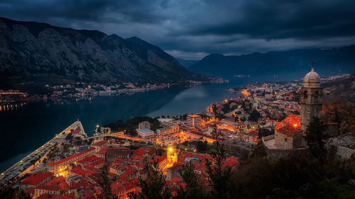 Una vista aerea di una città costiera illuminata con tetti rossi al tramonto, incastonata tra un'ampia baia e ripide montagne.