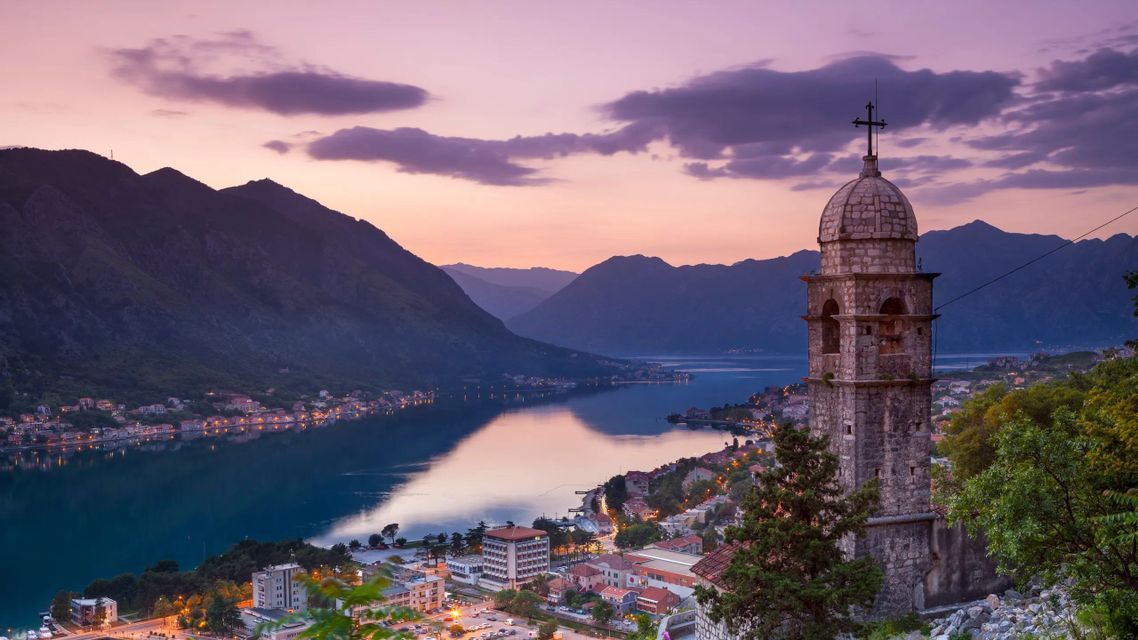 Al tramonto, un campanile di chiesa in pietra si affaccia su una cittadina costiera e una baia tranquilla circondata da montagne.