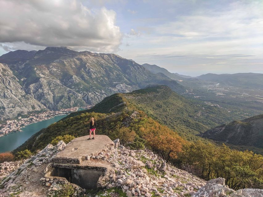 Una donna si trova su un bunker di cemento che si affaccia su un paesaggio montuoso, una città costiera e una baia sotto un cielo nuvoloso.