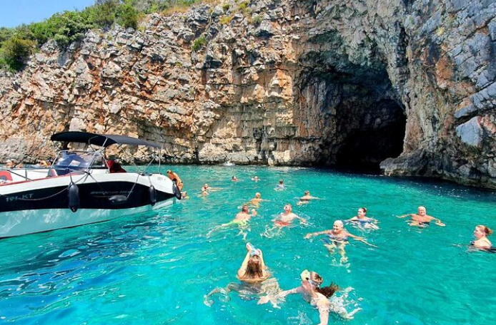 Un viaggio di gruppo WeRoad che nuota in acqua turchese accanto a un motoscafo, vicino all'ingresso di una grande grotta marina.