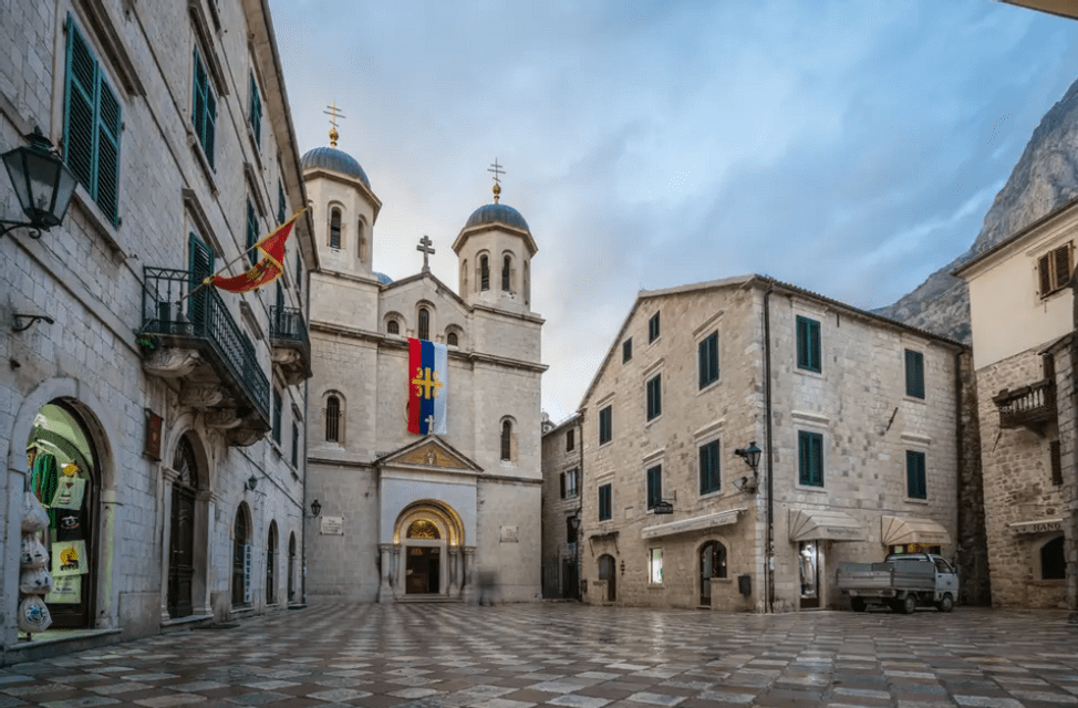 Una cattedrale in pietra con doppie cupole si erge in una piazza a scacchiera, affiancata da edifici storici e una montagna sullo sfondo.