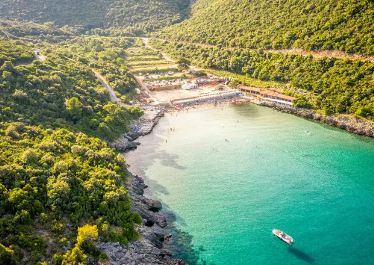 Veduta aerea di una piccola imbarcazione in una cala dalle acque turchesi, con una spiaggia incastonata tra scogliere rocciose e colline verdi.