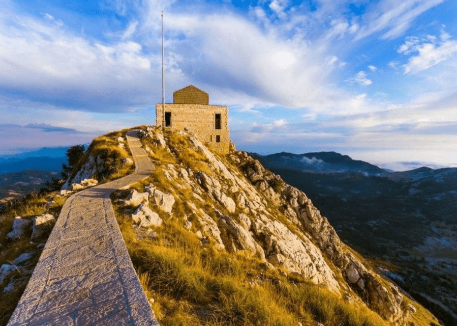 Un sentiero in pietra si inerpica su una montagna erbosa e rocciosa fino a un edificio di pietra sulla sua vetta, sotto un cielo azzurro con nuvole.