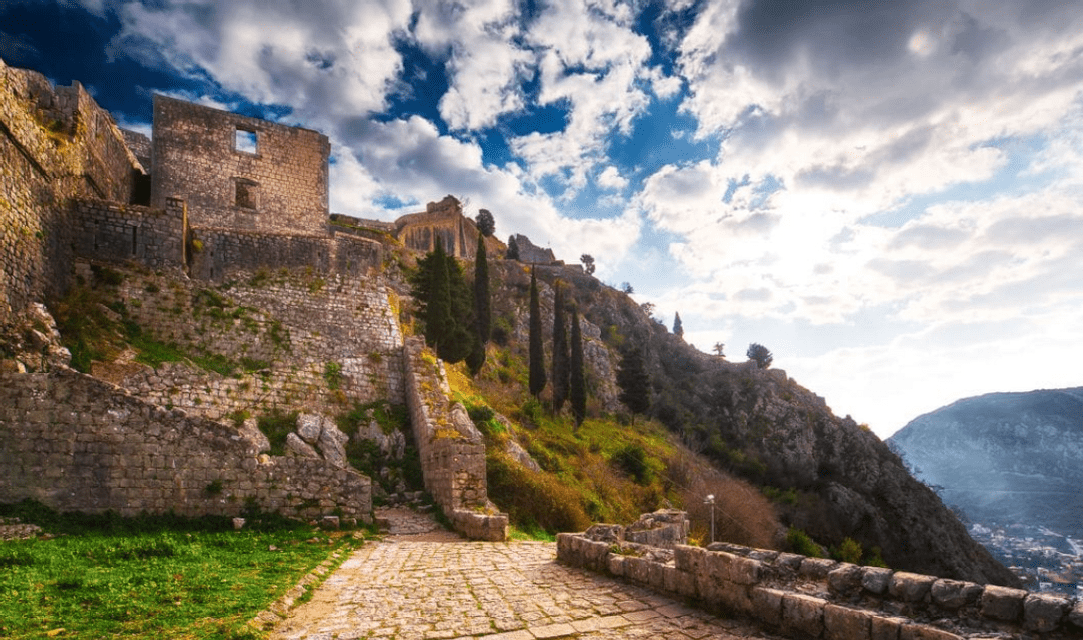 Rovine in pietra di una fortezza si ergono su un ripido pendio verde, con un sentiero di ciottoli che sale sotto un cielo nuvoloso.