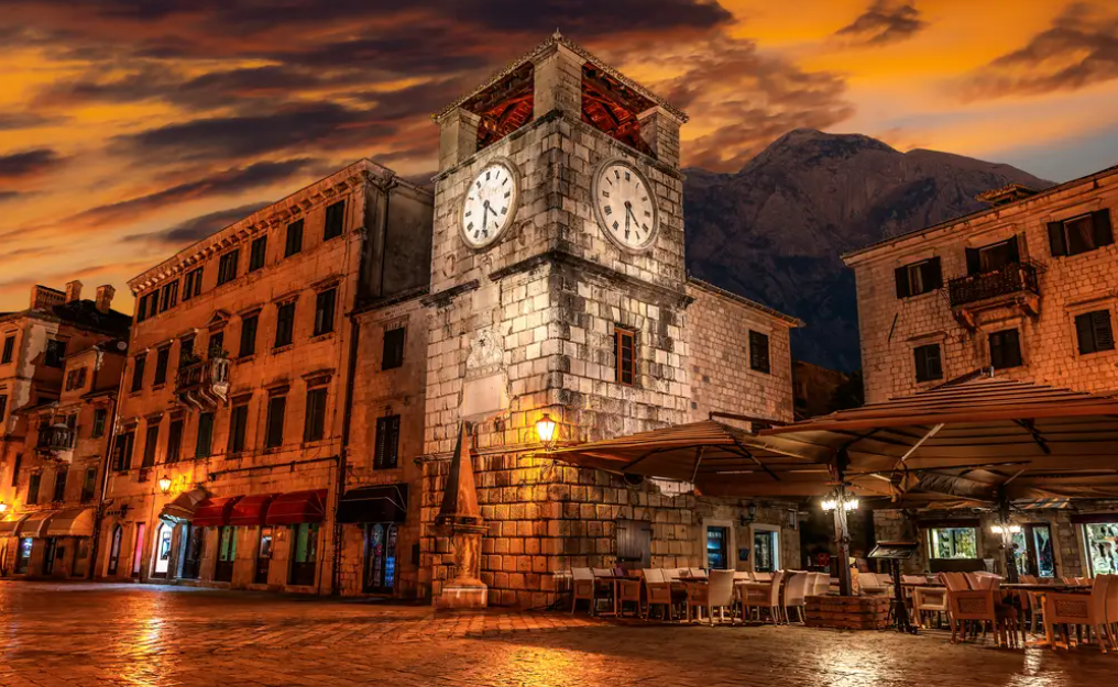 Una torre dell'orologio in pietra si staglia in una piazza deserta al tramonto, circondata da edifici illuminati e un caffè all'aperto.