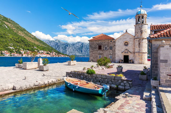 Un molo in pietra con una chiesa, con una piccola barca ormeggiata in acqua turchese limpida e montagne che si ergono sullo sfondo.