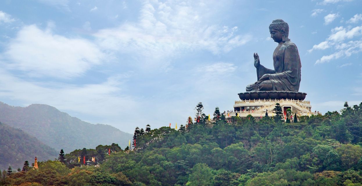 Une grande statue de Bouddha en bronze se dresse au sommet d'une colline boisée, sur une plate-forme à plusieurs niveaux, sous un ciel partiellement nuageux.