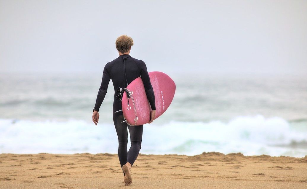 Un surfeur en combinaison noire tenant une planche de surf rose marche sur une plage de sable en direction de l'océan.