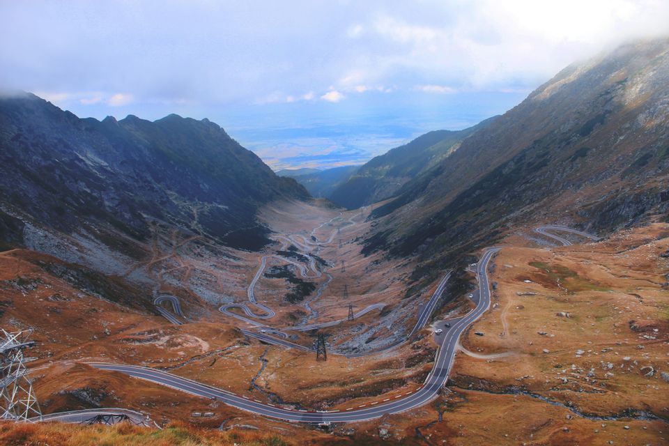Eine Luftaufnahme einer kurvenreichen Straße mit vielen Serpentinen, die sich durch ein großes Gebirgstal unter bewölktem Himmel windet.