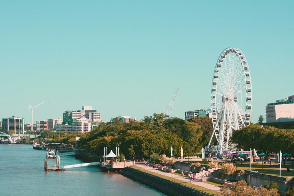 Une grande roue blanche se dresse sur les berges d'une ville, avec des gens se promenant sur une promenade bordée d'arbres verts luxuriants.