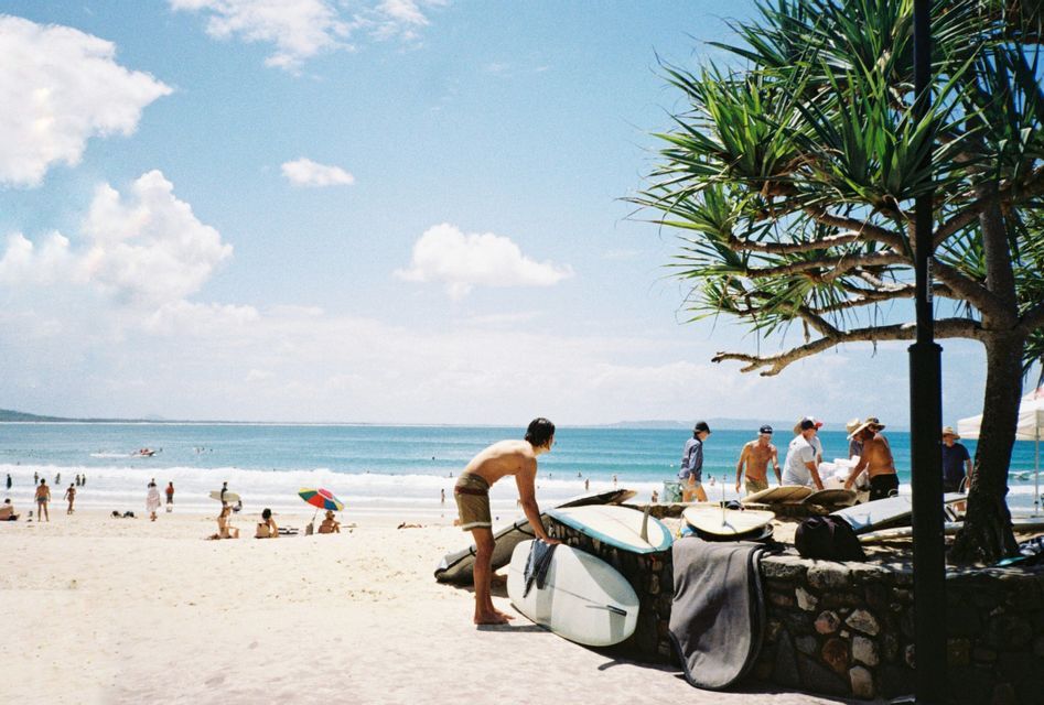 Un surfeur prépare sa planche sur un mur de pierre sur une plage animée, avec l'océan et d'autres personnes en arrière-plan.