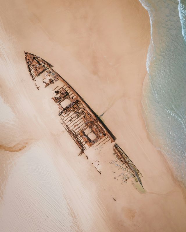 Vue aérienne d'une grande épave rouillée échouée sur le sable, avec de douces vagues de l'océan qui viennent s'échouer.