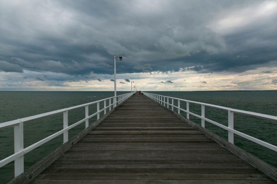 Une longue jetée en bois avec des balustrades blanches s'étend sur une mer sombre sous un ciel couvert et nuageux.