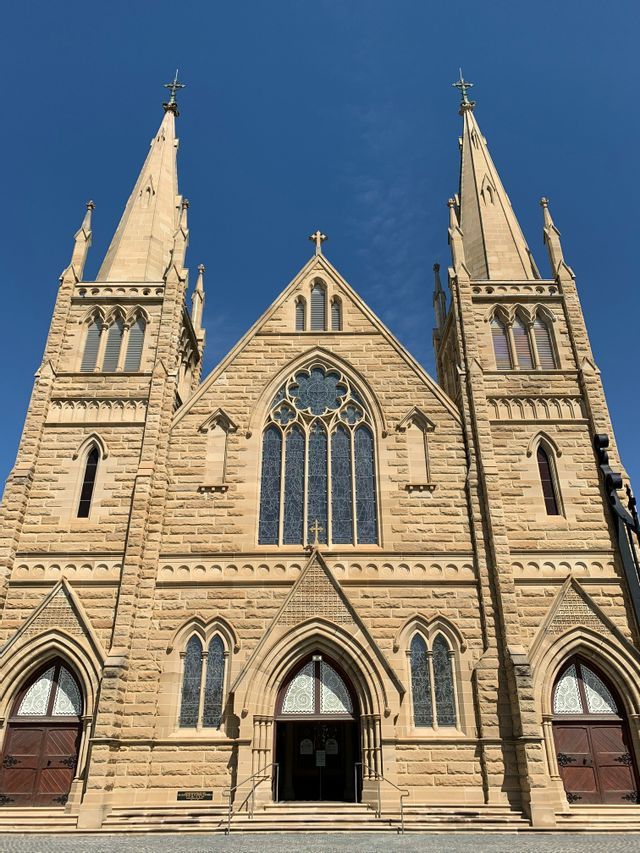 La façade d'une grande église en pierre avec deux flèches proéminentes, des arcs brisés et des vitraux sur un ciel bleu clair.