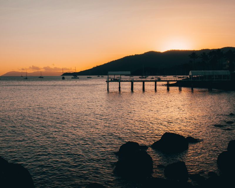 Le soleil se couche derrière une colline en silhouette, projetant une lueur orange sur une baie avec une jetée et des voiliers ancrés.