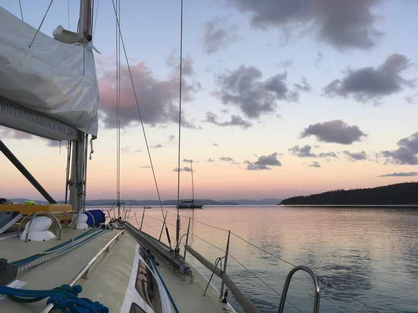 Vue depuis le pont d'un voilier sur une eau calme au coucher du soleil, avec des îles lointaines et d'autres bateaux à l'horizon.