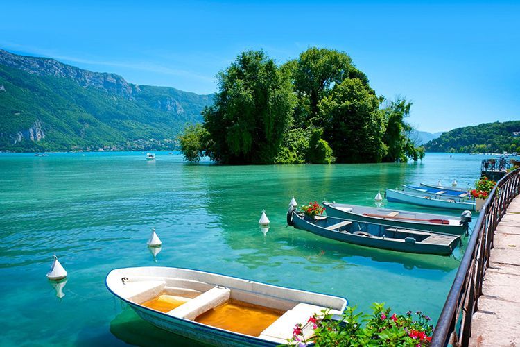 Barche a remi ormeggiate lungo una passeggiata su un lago turchese, con un'isola boscosa e montagne sullo sfondo.