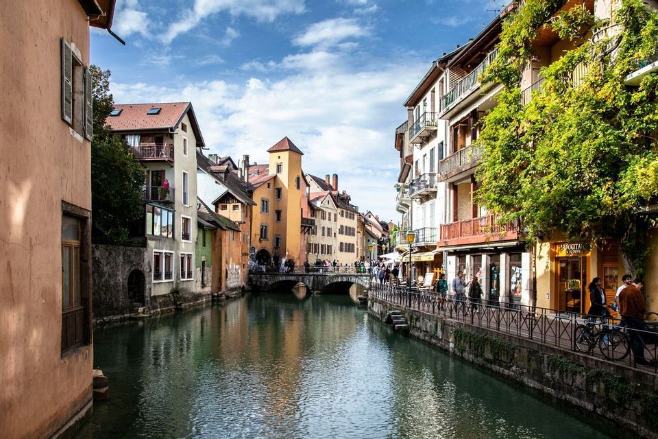 Un canale attraversa una città storica con edifici colorati, mentre le persone passeggiano lungo le rive e su un ponte di pietra.