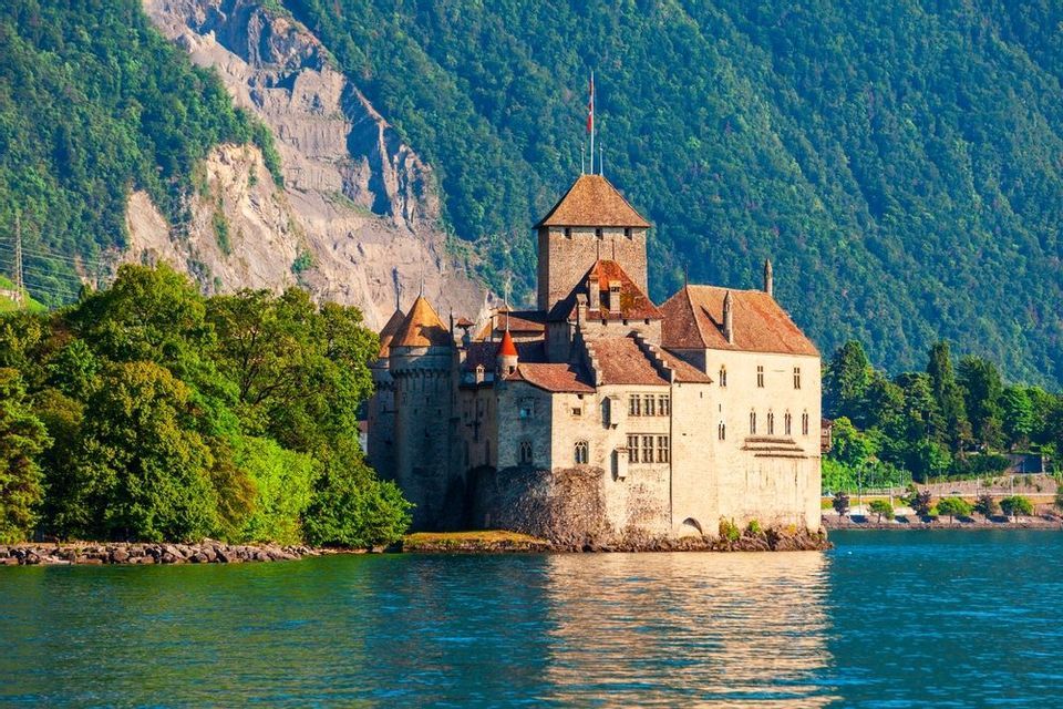 Un castello di pietra con tetti in tegole si trova sulla riva di un lago blu, con una montagna ripida e alberata sullo sfondo.