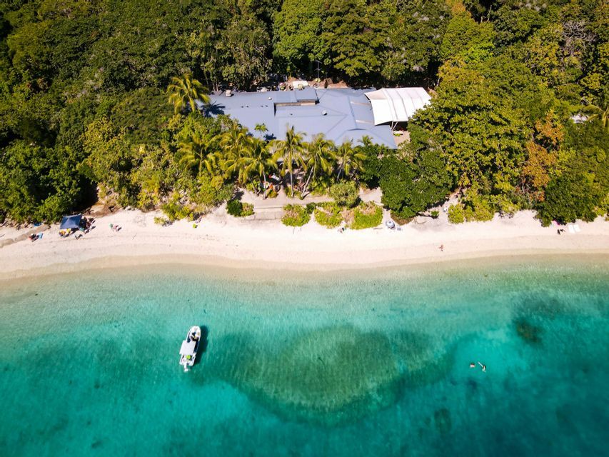 Une vue aérienne d'un littoral tropical avec une plage de sable blanc, une eau turquoise, un bateau et un bâtiment caché dans la forêt.