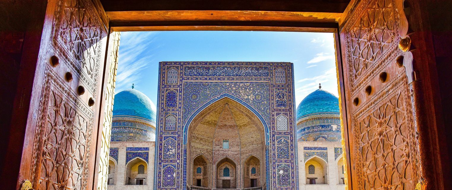 Una moschea ornata con cupole blu e un grande ingresso ad arco, vista attraverso due porte di legno aperte e finemente intagliate.