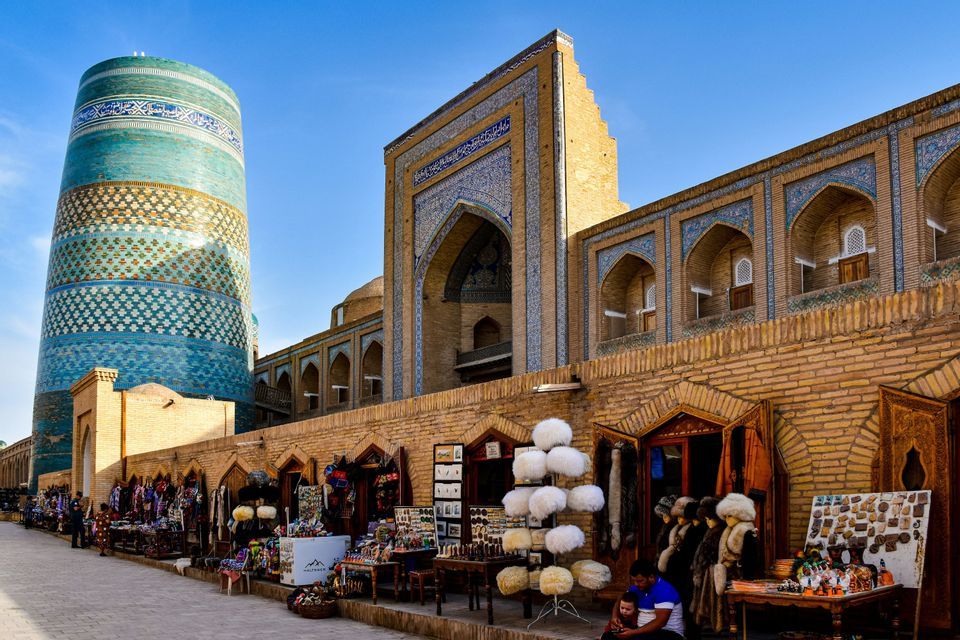 Des étals de marché en plein air longent un mur de briques, devant un minaret massif aux carreaux bleus et un bâtiment orné.