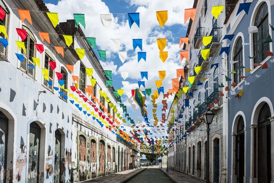 Colorful festival flags and a paper lantern hang over a narrow cobblestone street between old buildings under a partly cloudy sky.