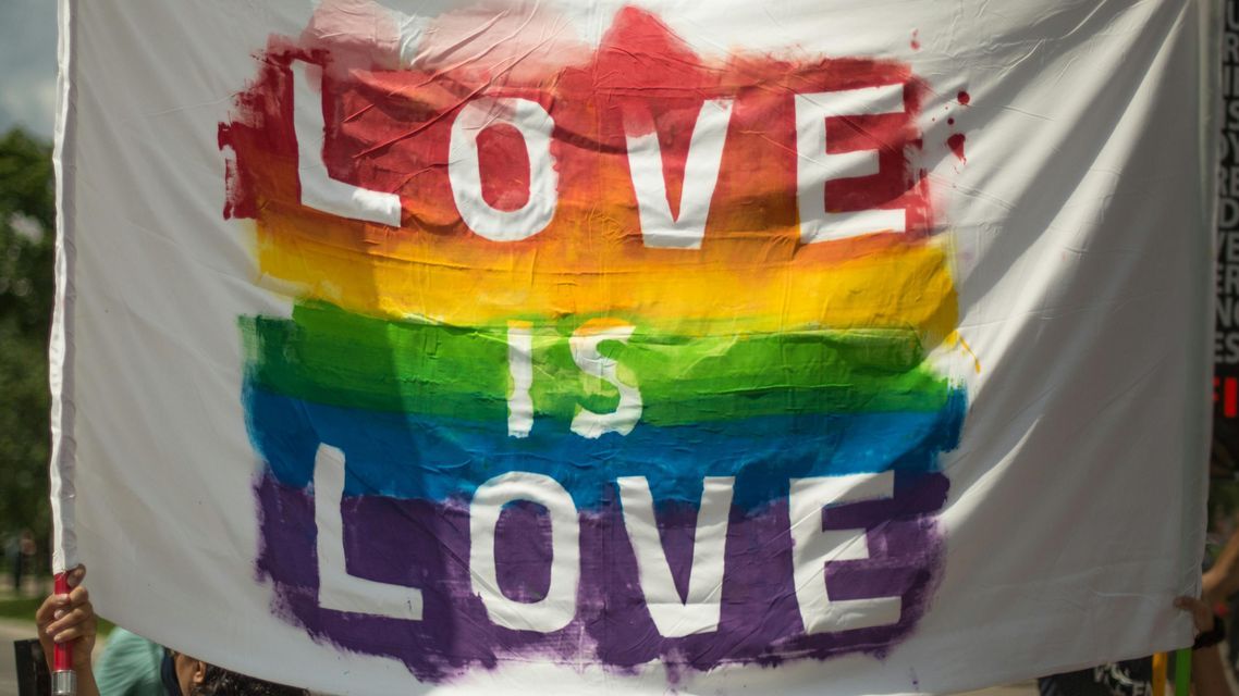 Uno striscione bianco con 'L'AMORE È AMORE' a caratteri cubitali su uno sfondo di colori arcobaleno dipinti a mano