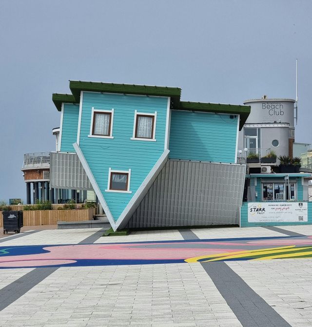 Una casa color verde acqua a due piani, costruita capovolta, che poggia sul tetto in una piazza pavimentata con disegni colorati sul terreno.