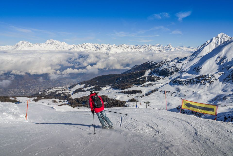 Ein Skifahrer in roter Jacke fährt einen schneebedeckten Berghang hinab, mit Blick auf ein Wolkenmeer und entfernte Gipfel unter einem klaren blauen Himmel.