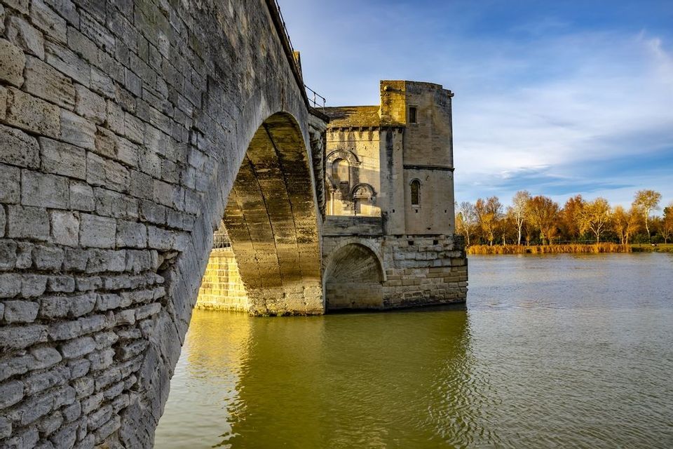 Eine historische Steinbrücke mit einer auf einem Pfeiler errichteten Kapelle überspannt einen breiten Fluss unter einem blauen Himmel mit Wolken.