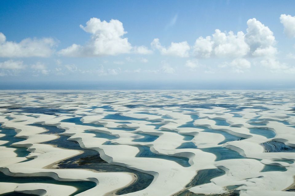 An aerial view of vast white sand dunes filled with lagoons of blue water under a partly cloudy sky.