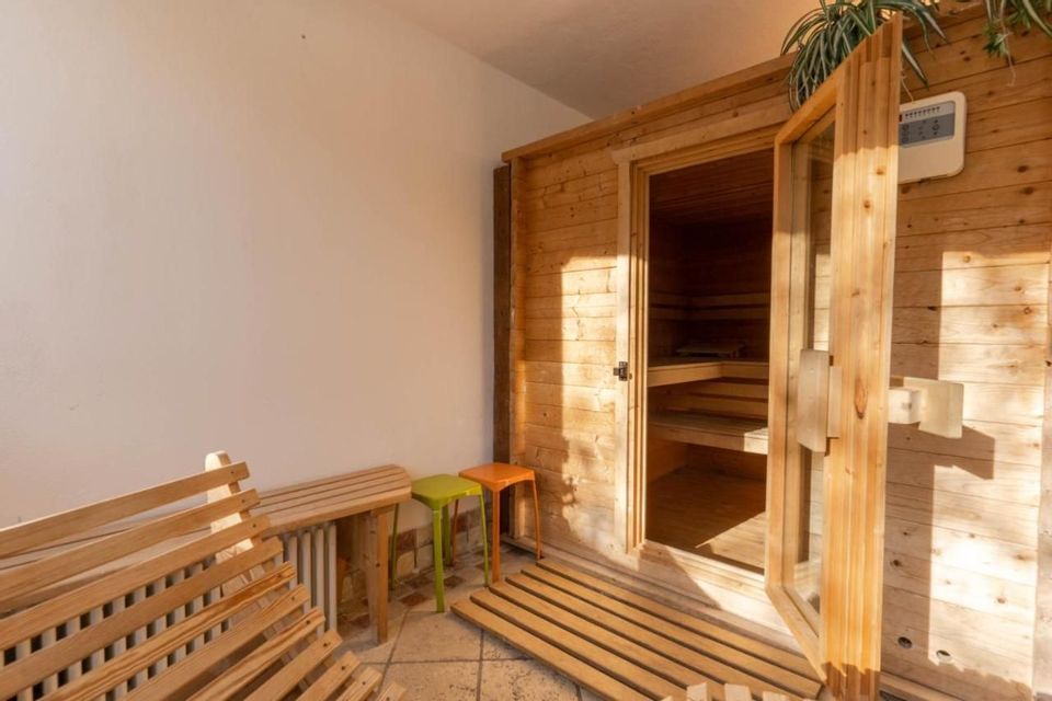 Una sauna in legno con porta a vetri aperta, situata in una stanza illuminata dal sole con panche e sgabelli.