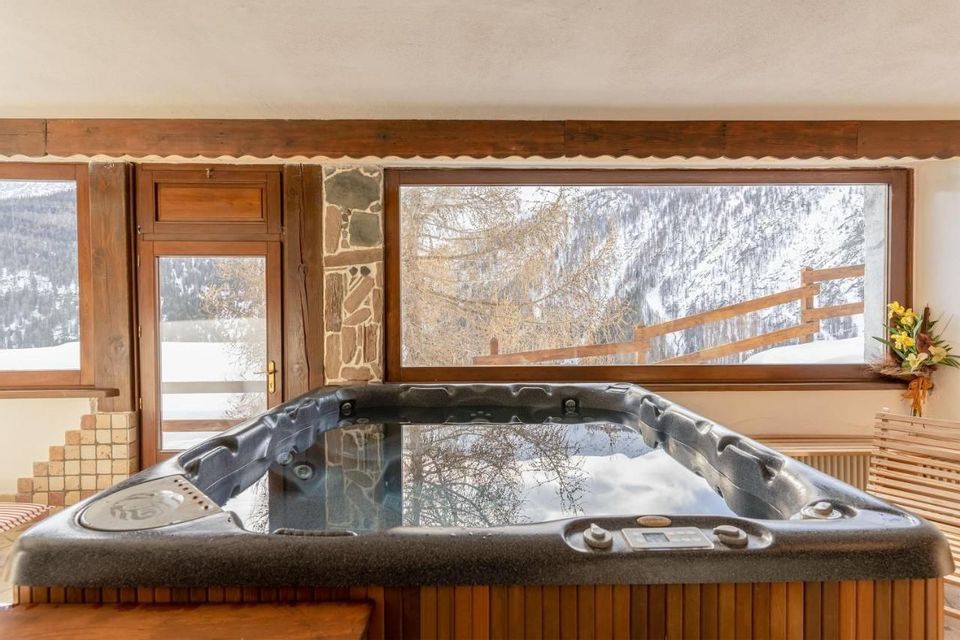 Vasca idromassaggio interna in camera rustica con ampia finestra su paesaggio montano innevato.