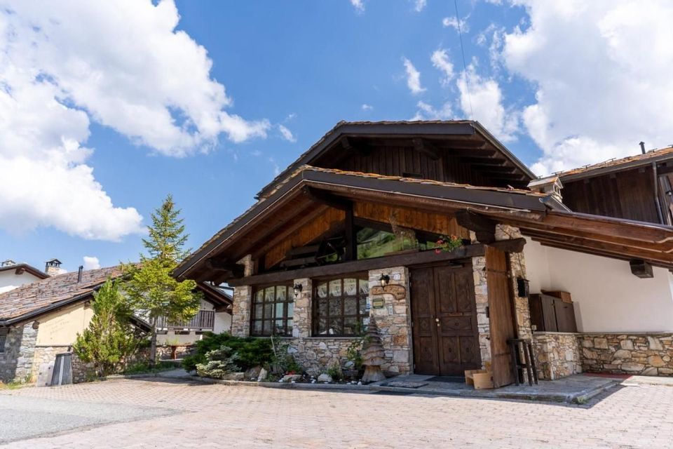 Un edificio rustico in stile chalet, con una base in pietra e un piano superiore in legno, si staglia contro un cielo azzurro con nuvole bianche.
