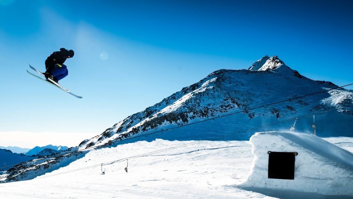 Uno sciatore è a mezz'aria mentre esegue un salto su una montagna innevata con un cielo azzurro limpido sullo sfondo.