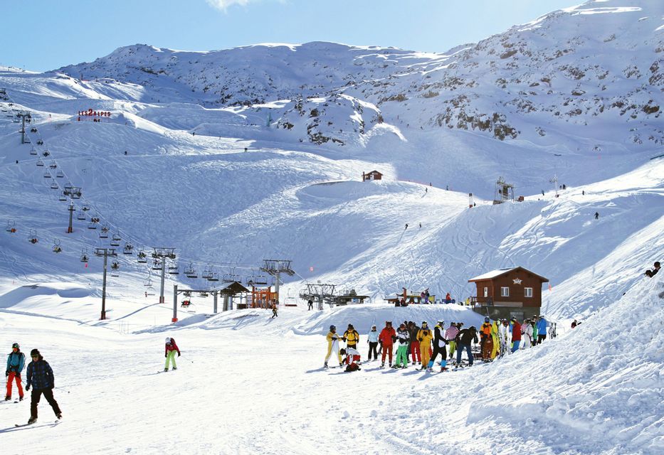 Un viaggio di gruppo WeRoad con attrezzatura da sci colorata su una montagna soleggiata e innevata, con seggiovie e altri sciatori sullo sfondo.