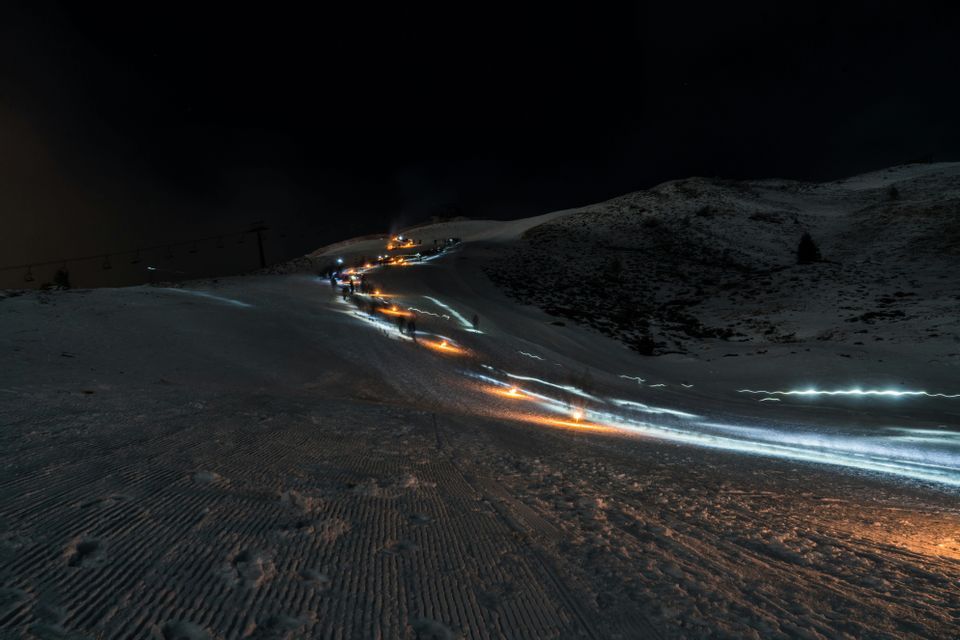 Uno scatto a lunga esposizione di un gruppo WeRoad in viaggio che sale un pendio innevato di notte, con le torce che creano scie di luce arancioni e bianche.