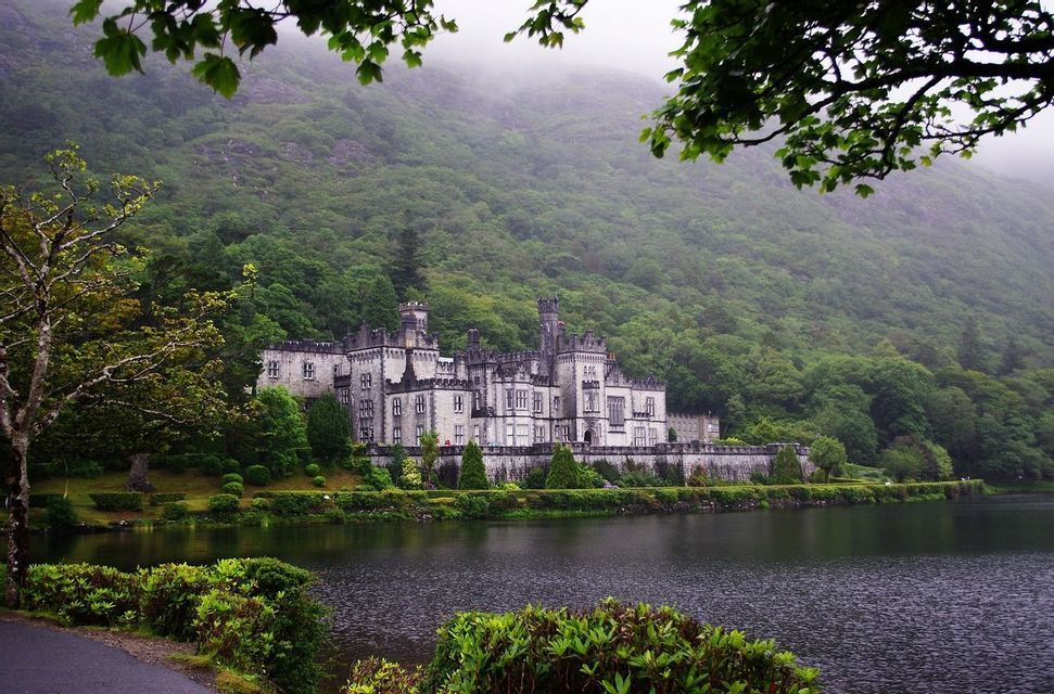 Un grand château de pierre se dresse sur les rives d'un lac, avec une colline escarpée et densément boisée s'élevant dans la brume en arrière-plan.