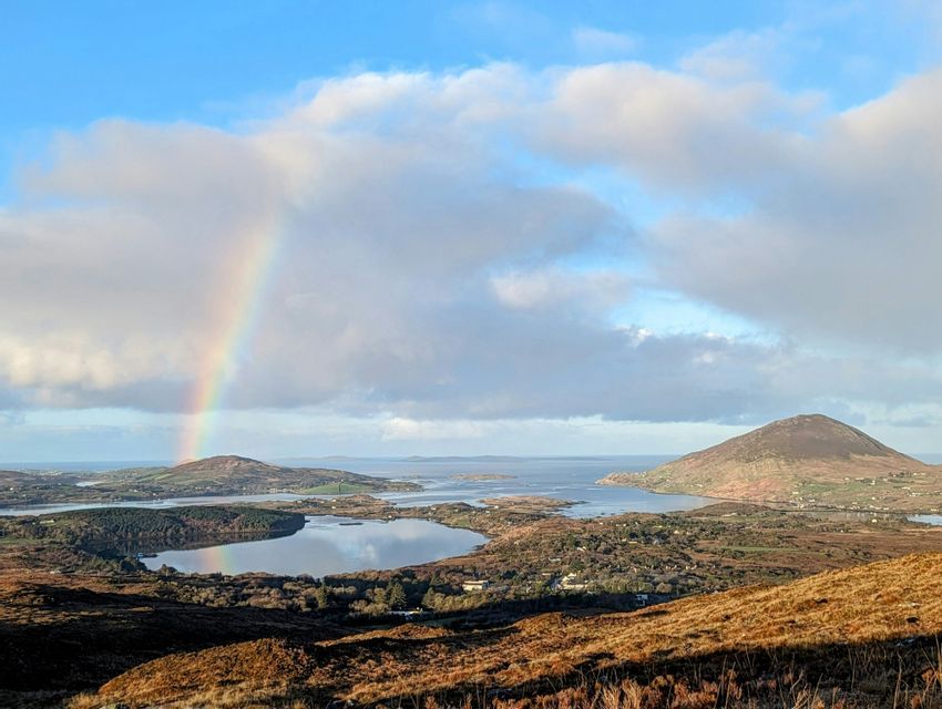 Un arc-en-ciel se forme au-dessus d'un paysage côtier de collines et d'un lac, avec son reflet visible à la surface de l'eau.